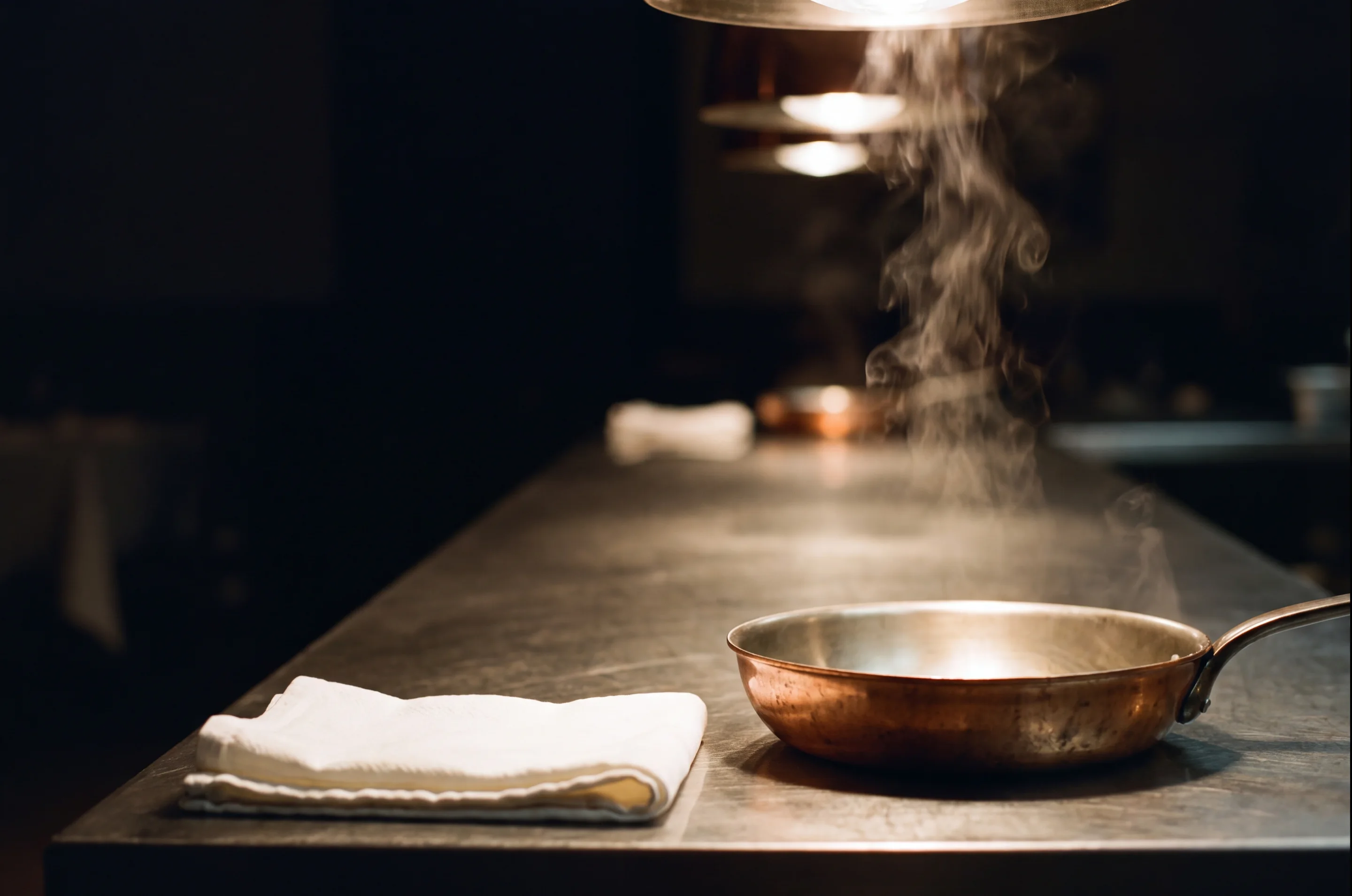 A professional kitchen pass in the last quiet minute before service: brushed steel counter, heat lamp, column of steam, folded linen, copper pan. A generated image of the trade — no identifiable venue, no campaign, no delivered work.